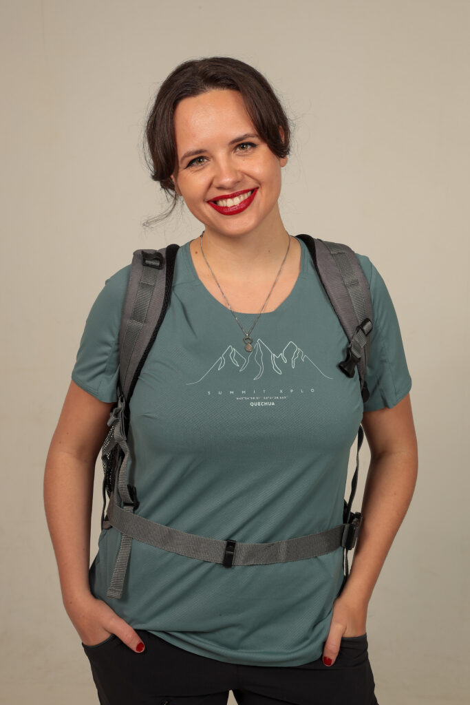 Marie Elise, zij heeft bruin haar en staat breed glimlachend, staande met haar handen in haar zakken en een rugzak om.